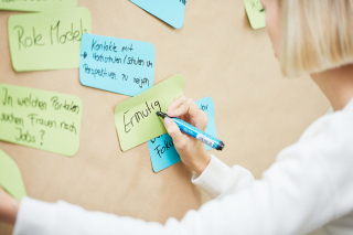 Eine blonde Frau schreibt Schlagwörter auf farbige Kärtchen, die auf einer Pinnwand befestigt sind.